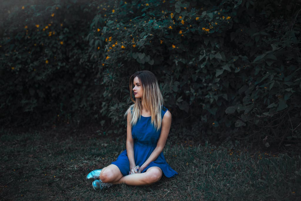 A young woman in a blue dress sits on the grass surrounded by lush foliage.