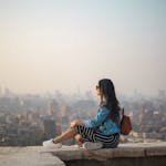 A young woman in casual attire sits on a rooftop terrace, viewing a sprawling cityscape in daylight.