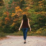 A young woman walks down a colorful autumn forest path, surrounded by vibrant leaves.